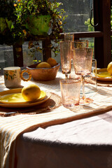 A bright summer table decorated with lemons, green hydrangeas, and glassware. Sunlight pours through hanging plants, creating a cozy, cheerful atmosphere perfect for outdoor dining