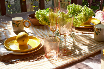 A bright summer table decorated with lemons, green hydrangeas, and glassware. Sunlight pours through hanging plants, creating a cozy, cheerful atmosphere perfect for outdoor dining