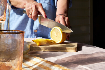 Close-up of a woman slicing a lemon on a wooden board. Sunlight highlights fresh citrus, glassware,...
