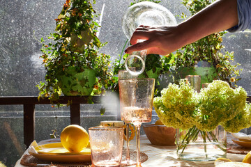 Close-up of a hand pouring water into a glass on a sunlit summer table. Green hydrangeas, lemons,...