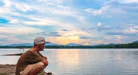 An elderly Asian man sits by the water in the evening.