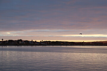 The sun rises above a calm bay, casting a golden glow over wooden picnic tables and tranquil...