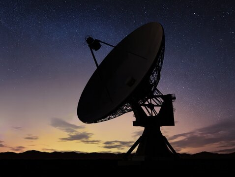 A prominent parabolic satellite dish is silhouetted during twilight, surrounded by vast starry skies