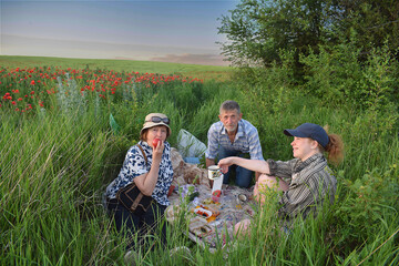 Elderly father, mother and adult daughter on a picnic in the steppe among blooming poppies.