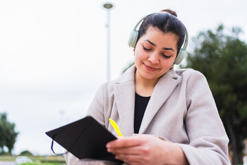 Young influencer writing notes on notebook while listening to music with headphones sitting in a park