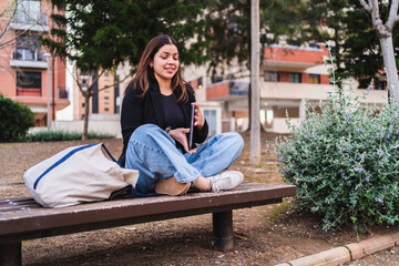 Young woman using digital tablet, creating online content while sitting on bench in city park