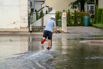 Boy riding scooter through urban floodwater in summer – fun, childhood and weather concept