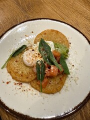 A dish served on a white plate with crispy rounds, poached egg, avocado, diced tomatoes, grated cheese, spices, and fresh green leaves on top.