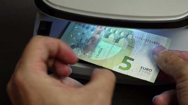Examination of five Euro (EUR) cash money with UV counterfeit detector. Concept of authenticity check, fight against counterfeiting of banknotes, anti fraud measures in European Union. Close up view.
