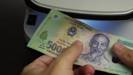 Examination of vietnamese dong (VND) cash money with UV counterfeit detector. Concept of authenticity check, fight against counterfeiting of banknotes, anti fraud measures in Vietnam. Close up view.