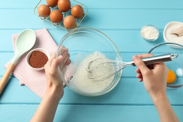 Woman whisking egg whites in bowl at light blue wooden table, top view