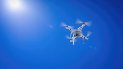 A white drone is seen flying steadily in a bright blue sky filled with sunlight. The clear weather emphasizes the drone's sleek design, showcasing its rotors in motion