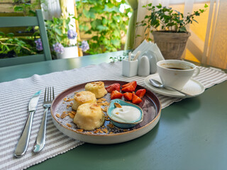 Gourmet breakfast served outdoors: A plate of syrniki with fresh strawberries on a green wooden table.