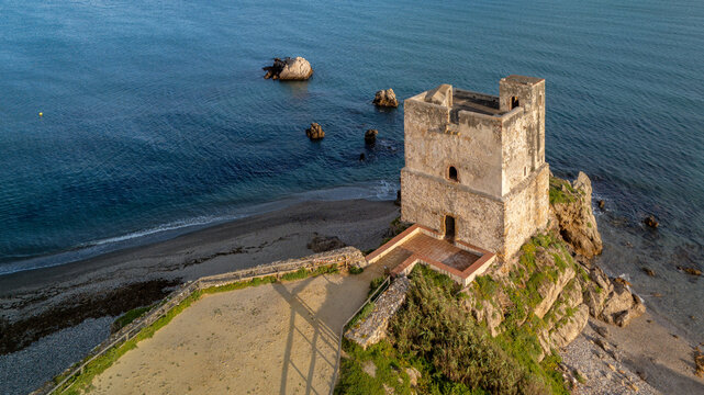 Casares Coastal Watchtower: An ancient watchtower stands on a rocky outcrop at the water's edge, with a sandy beach and a town of white buildings dotting the distant hills under a cloudy sky - Powered by Adobe