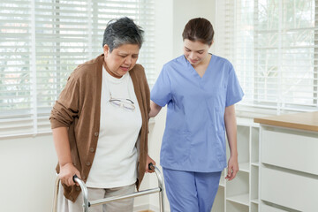 Senior asian female patient walking slowly with walker under supervision of female physiotherapist...