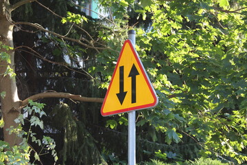 Yellow triangular warning road sign with exclamation mark mounted on metal pole surrounded by green...