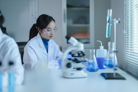 Asian female scientist using microscope analyzing sample in laboratory glassware, conducting scientific experiment research