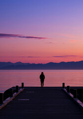 Fototapeta premium Silhouette of a person standing on a pier watching a beautiful pink and purple sunset over the ocean and mountains