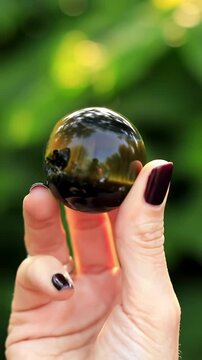 An extreme close-up, slow-motion video captures a fair-skinned hand, adorned with silver rings and dark nail polish, rotating a polished tiger's eye stone sphere, its chatoyancy shimmering with
