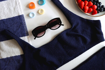 Navy swimsuit, striped beach towel, sunglasses, colorful rings and plate with berries. Top view.