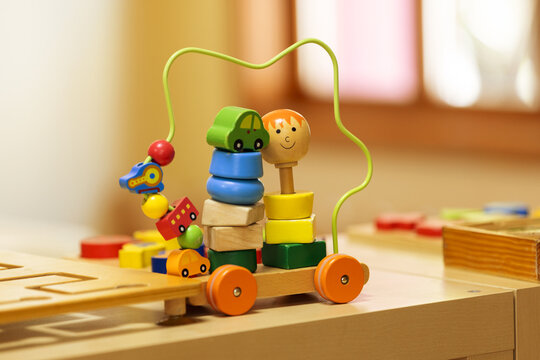 A colorful wooden bead maze and stacking toy with wheels sits on a light wooden surface in a kindergarten setting. The image highlights engaging educational play for young children.