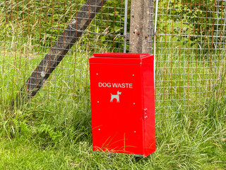 Red metal bin for dog waste attached to a fence in a publlic park. No people.