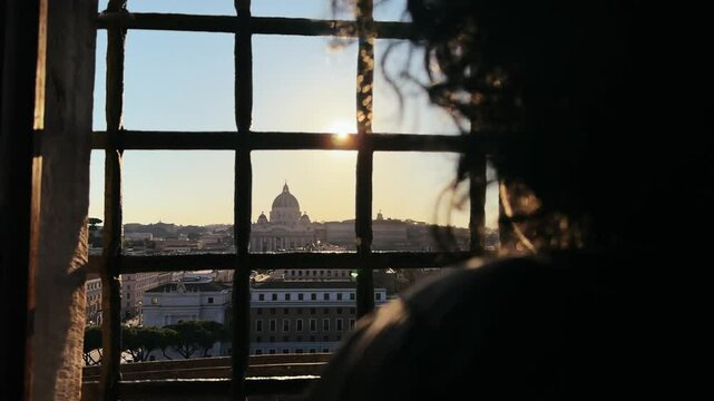 Silhouette of a person gazing out through a barred window at a cityscape of Rome with view of Saint Peter basilica at background seen from castle Castel Sant'Angelo
