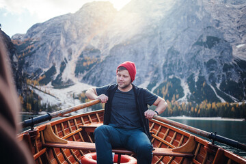 A man in a red beanie sits in a wooden rowboat on a clear lake, surrounded by majestic mountains...