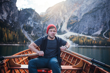 A man in a red beanie sits in a wooden rowboat on a clear lake, surrounded by majestic mountains...