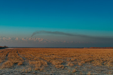 Winter field and mountains landscape with smoke from high pipe in the distance, Kyrgyzstan