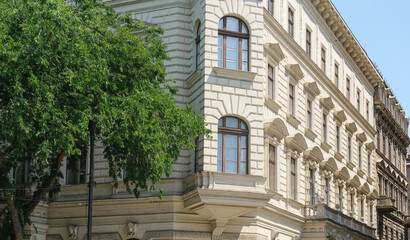 A corner view of a classic building with detailed stonework and large windows, surrounded by lush green trees.