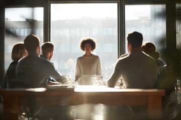 A woman presents to a small group seated around a long wooden table, bathed in sunlight streaming through a large window
