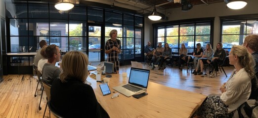 A woman presents to a small group seated in a modern, sunlit room with large windows and hardwood floors. A long wooden table holds laptops and tablets