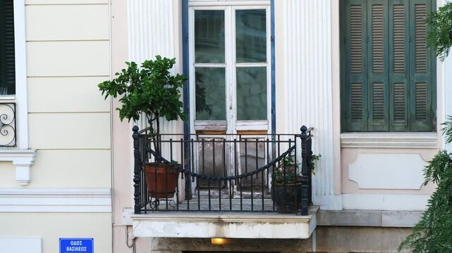 Detailed view of an old wrought iron balcony with potted plants, weathered doors, classical pilasters, and a street sign reading "Vasileos Street" in Athens, Greece 
