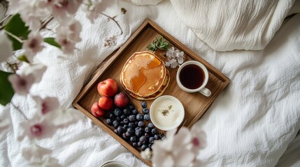 Overhead breakfast tray with pancakes, syrup, fresh fruit, and tea on white bedding