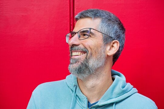 Portrait of a cheerful bearded man wearing eyeglasses and a light blue hoodie, smiling and looking away against a vibrant red background