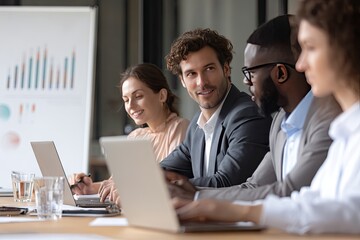 A diverse group of professionals collaborates around a table, using laptops and reviewing charts on a whiteboard during a meeting.  They appear engaged and positive