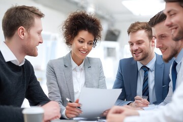 A diverse group of smiling business professionals are gathered around a table, reviewing documents together in a bright office setting
