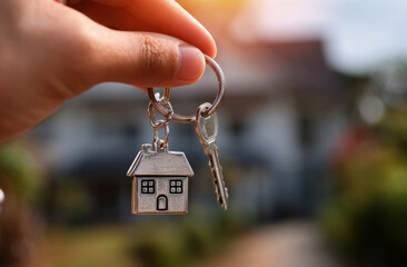  hand holding a silver house-shaped keychain with a blurred background of a home exterior on a sunny day
