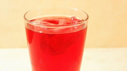 A tilted glass filled with vibrant red liquid sits against a soft peach background. The transparency of the drink and the gentle lighting create a clean and refreshing visual contrast.