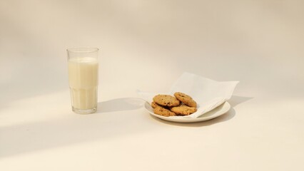 A glass of milk sits beside a plate of three cookies on a light-colored surface. A white cloth napkin is draped nearby. Soft natural light creates a cozy and inviting atmosphere.