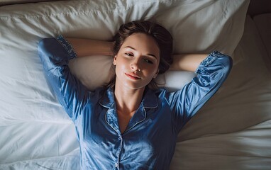 A young woman with brown hair lies supine on a bed in blue satin pajamas, hands behind her head, gazing serenely at the camera in soft, natural light