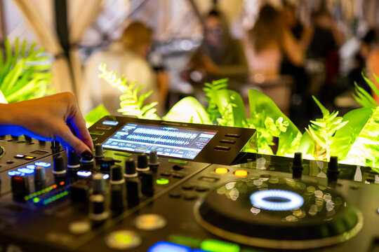 DJ mixing tracks on professional audio console with vibrant LED display, surrounded by greenery and blurred crowd, capturing dynamic party atmosphere.