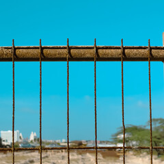 a weathered, rusty metal fence or barrier isolated against a clear blue sky. hints of buildings and...