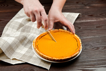 Hands of a baker slicing into a freshly baked pumpkin pie on a wooden table, with a checkered cloth beside it, showcasing the delicious dessert preparation process