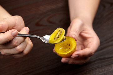 Close-up of hands holding a spoon with a sliced yellow kiwi fruit, showcasing its vibrant color and unique texture against a dark wooden background, emphasizing freshness and healthy eating