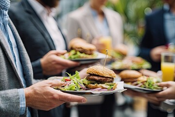 Business professionals informally gathered outdoors, holding plates with juicy burgers and side salads, enjoying a casual lunch break