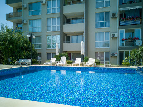 Clean outdoor pool with clear blue water, sun loungers, and umbrellas near modern apartment building. Peaceful summer vibe and relaxation area.