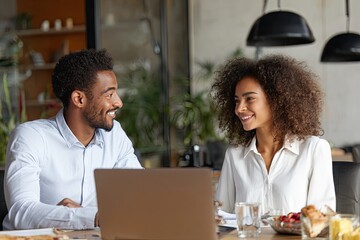 A smiling Black man and woman engage in conversation across a laptop at a wooden table in a cafe, enjoying a light meal and drinks