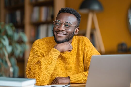 A smiling Black man in glasses, wearing a mustard yellow sweater, sits at a desk with a laptop, hand on chin, thoughtfully looking off to the side in a home library setting